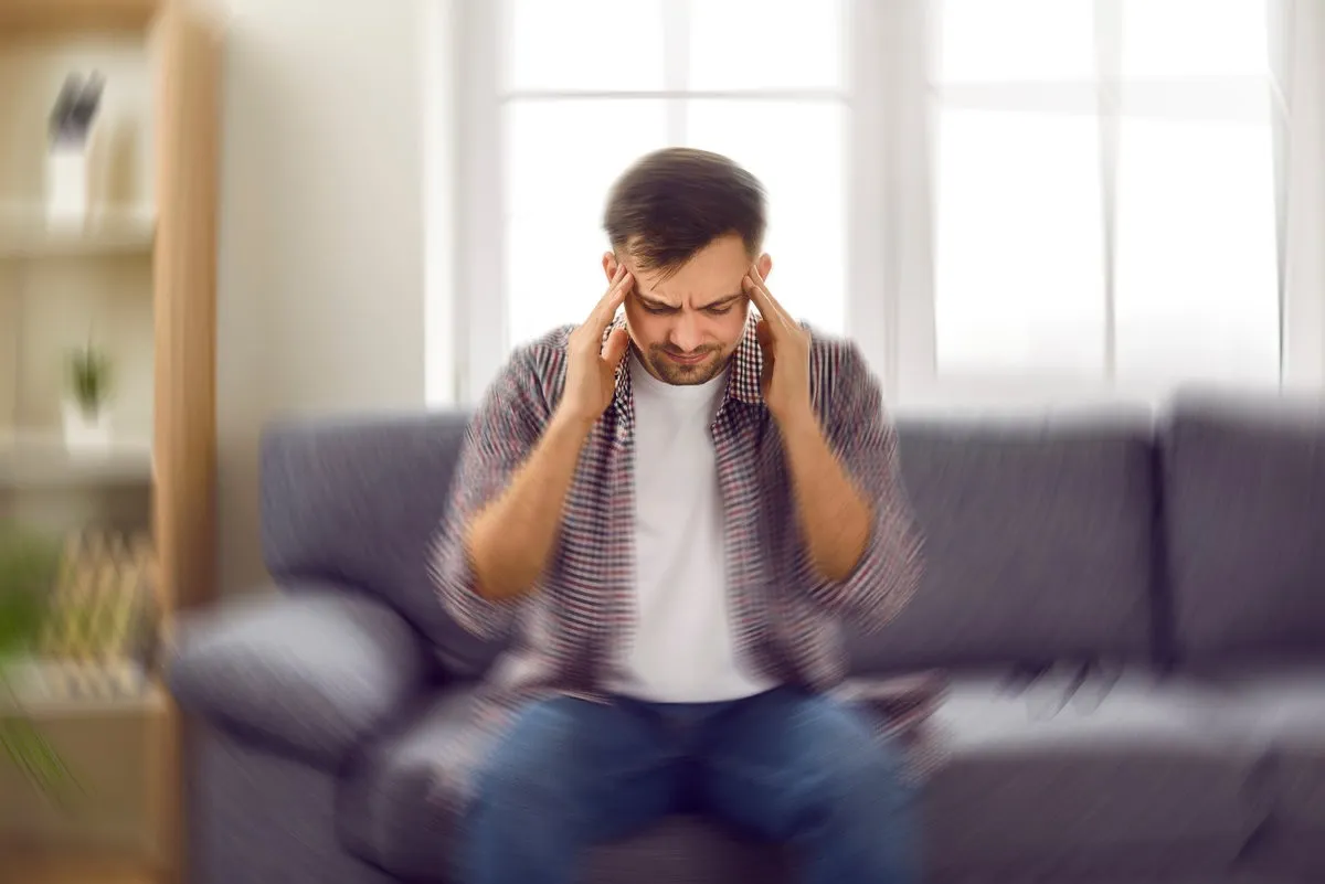 Man suddenly feels dizzy and takes a seat on the sofa. Young guy feeling pain and spinning sensation in his head.