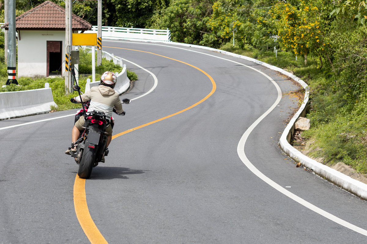 A lone motorcyclist navigates a winding asphalt road through lush green scenery. The road features clear white and yellow lines, leading into a series of curves typical of mountain roads.